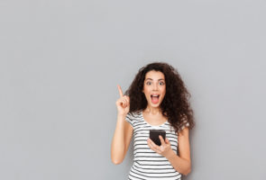 Smart female with mobile phone in hand keeping index finger pointed upwards expressing she has an idea or saying eureka, over grey background